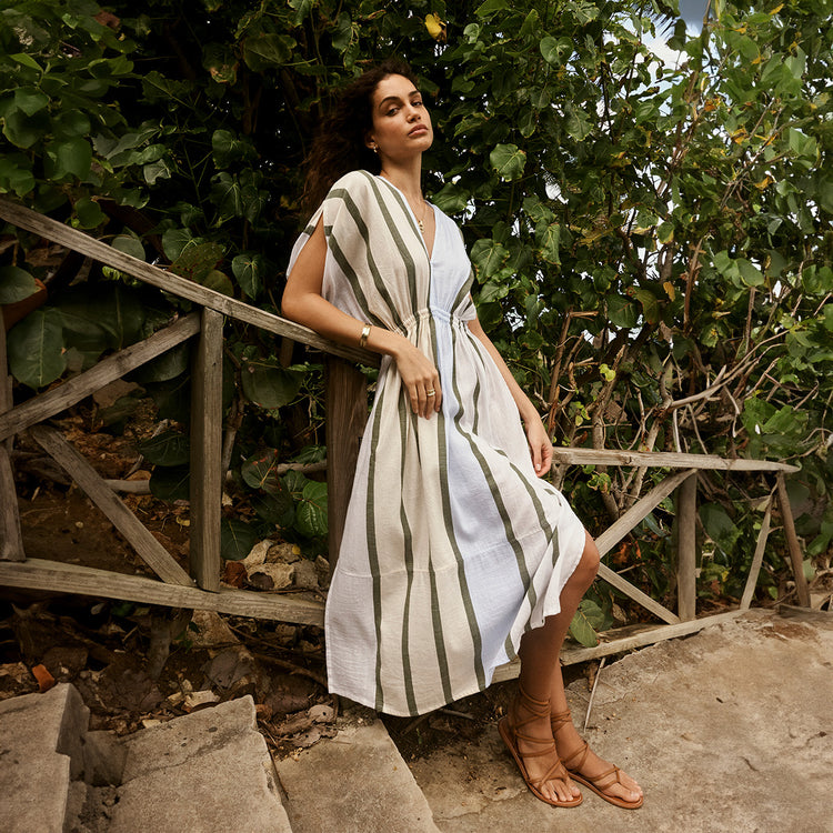 Woman standing against the wooden railing of stones stairs wearing a hand woven lemlem long plunge neck dress in white and light blue with sage stripes. 
