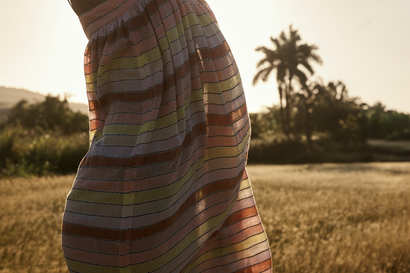close up on a woman body wearing a lemlem handwoven dress with pink, red, orange and yellow stries in a field in the carribeans. 