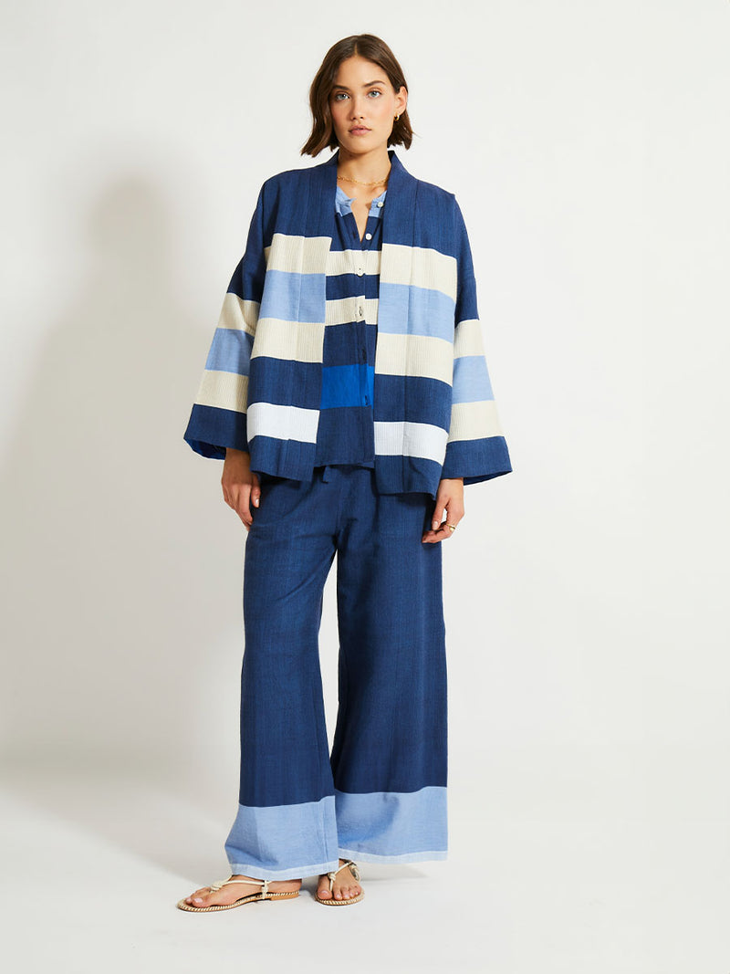 Woman Standing Wearing lemlem Desta Pants, Terrara Jacket and blouse featuring a striking color-block combination of three shades of blue, paired with bands of neutral cream and white, inspired by handmade indigo quilting and African textiles